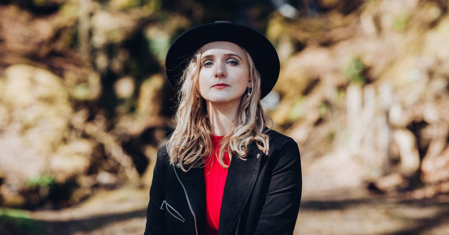 Kate Ferguson (Ethira) in a promotional image wearing red and black including a black hat