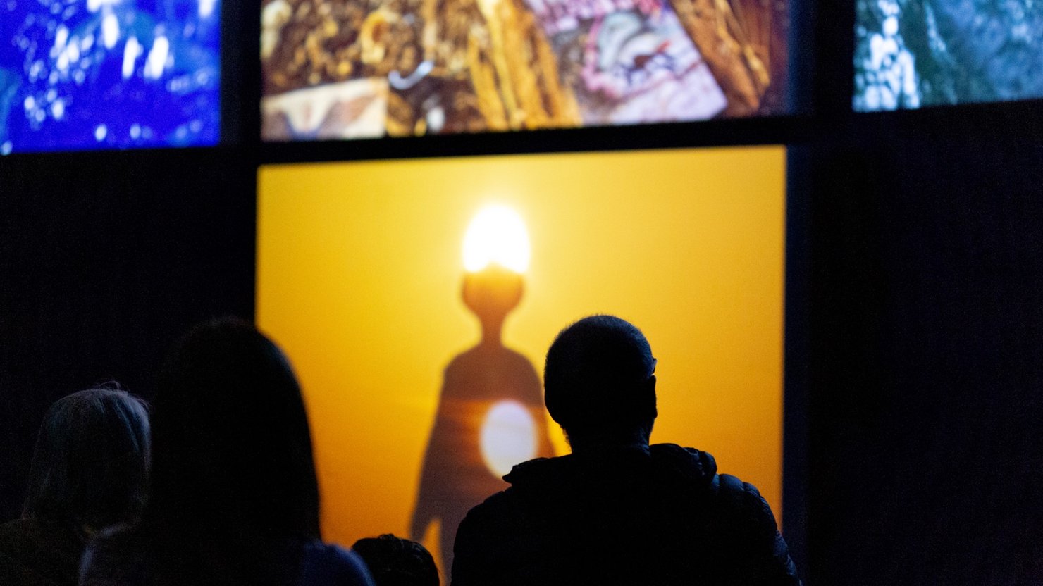 Audience watching film installation with screens showing a figure and sun