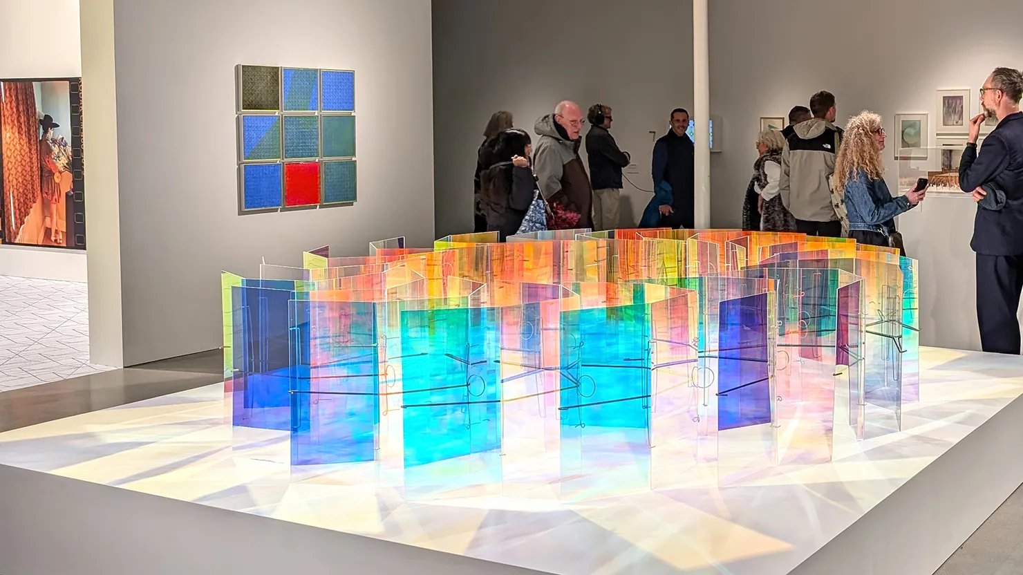 Photograph showing visitors looking at an exhibition with artwork on the walls and a large perspex installation in the foreground