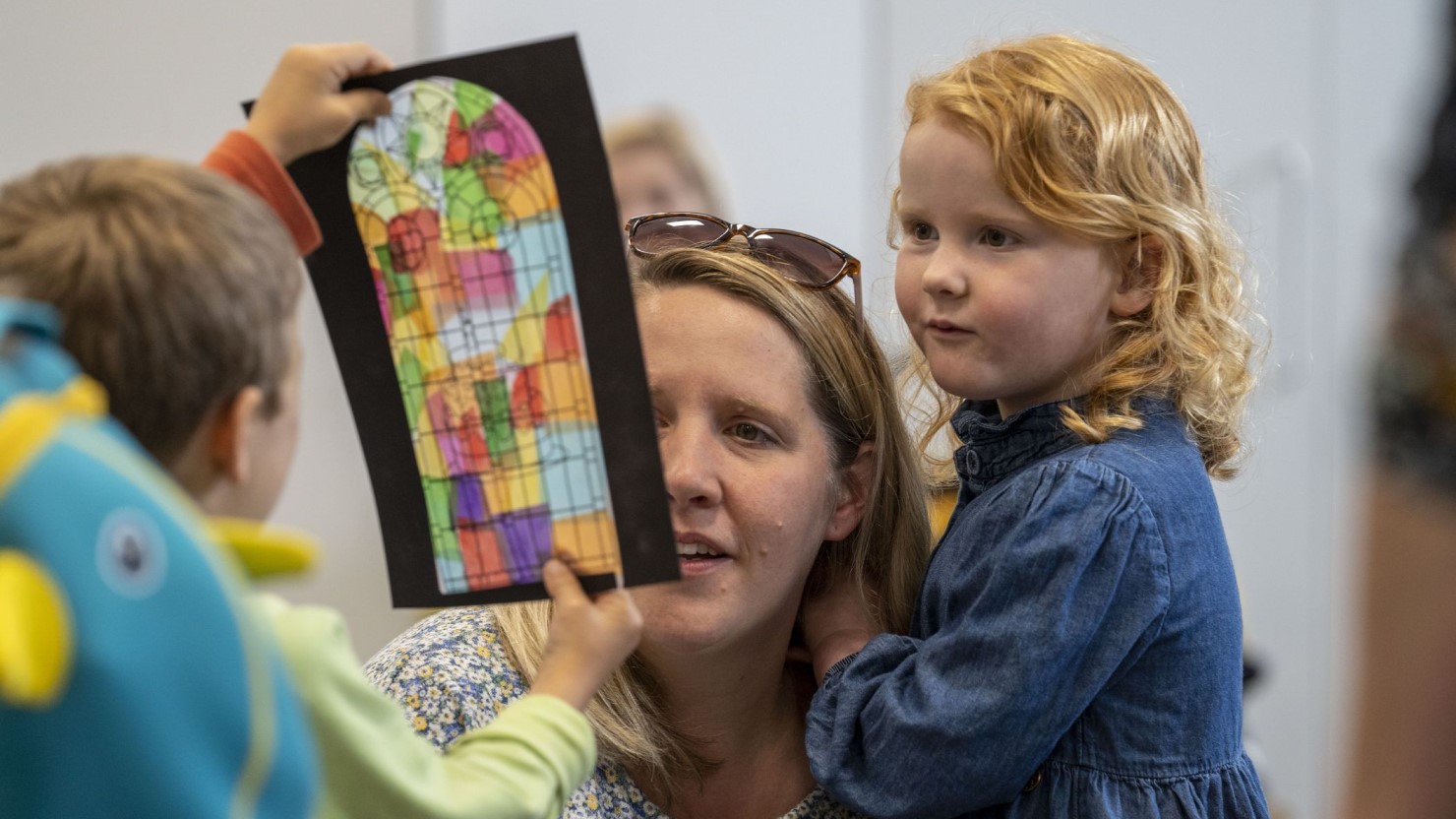 A boy showing a mum and daughter a stained glass artwork made in I Wonder workshop.