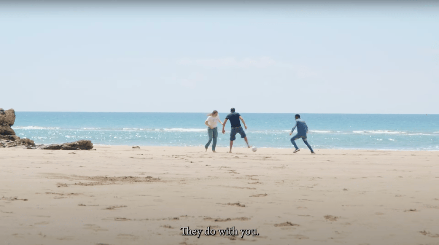 Still from a film showing three people playing football on a beach and a caption 'They do with you'.