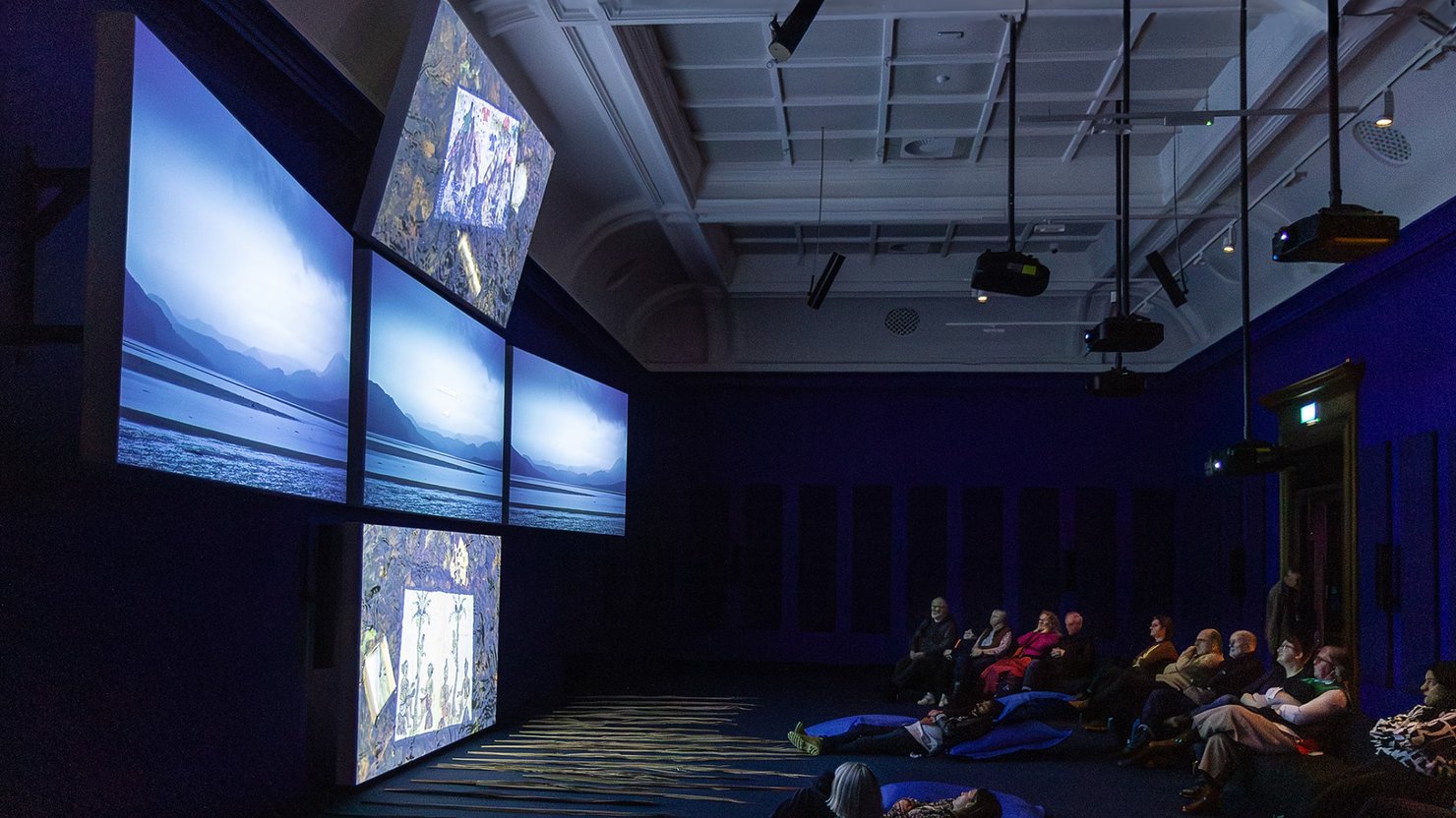 Audience sitting in a gallery watching film installation on five screens in a crucifix shape