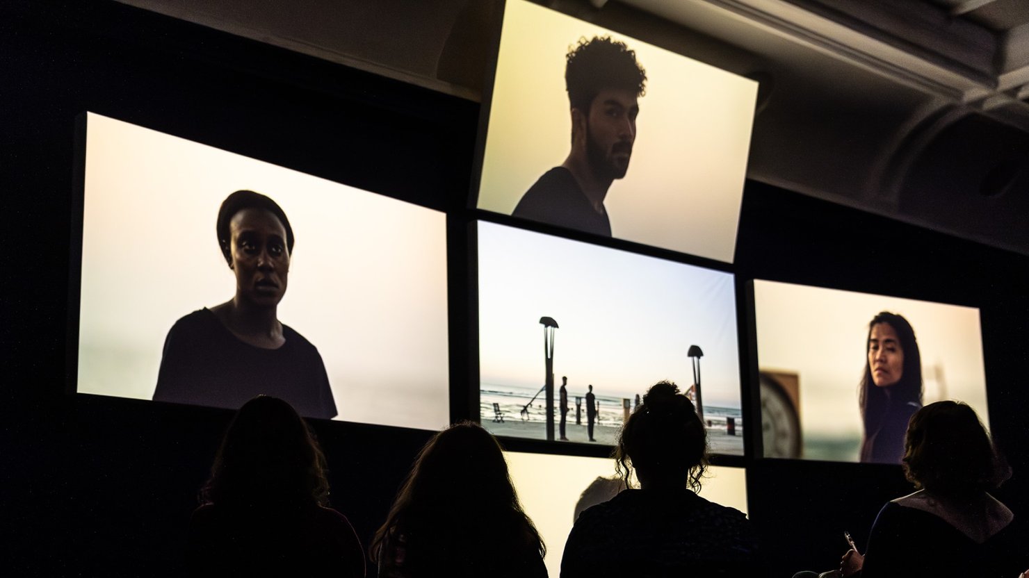 Audience watching film installation with screens showing people standing on a beach