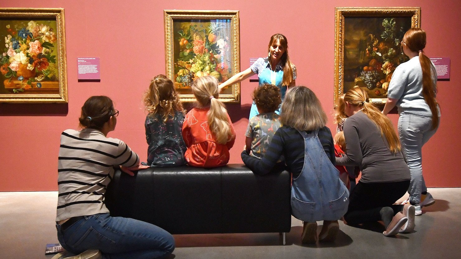 A group of children and adults sit on a black bench as they look at a flower painting hanging on a gallery wall