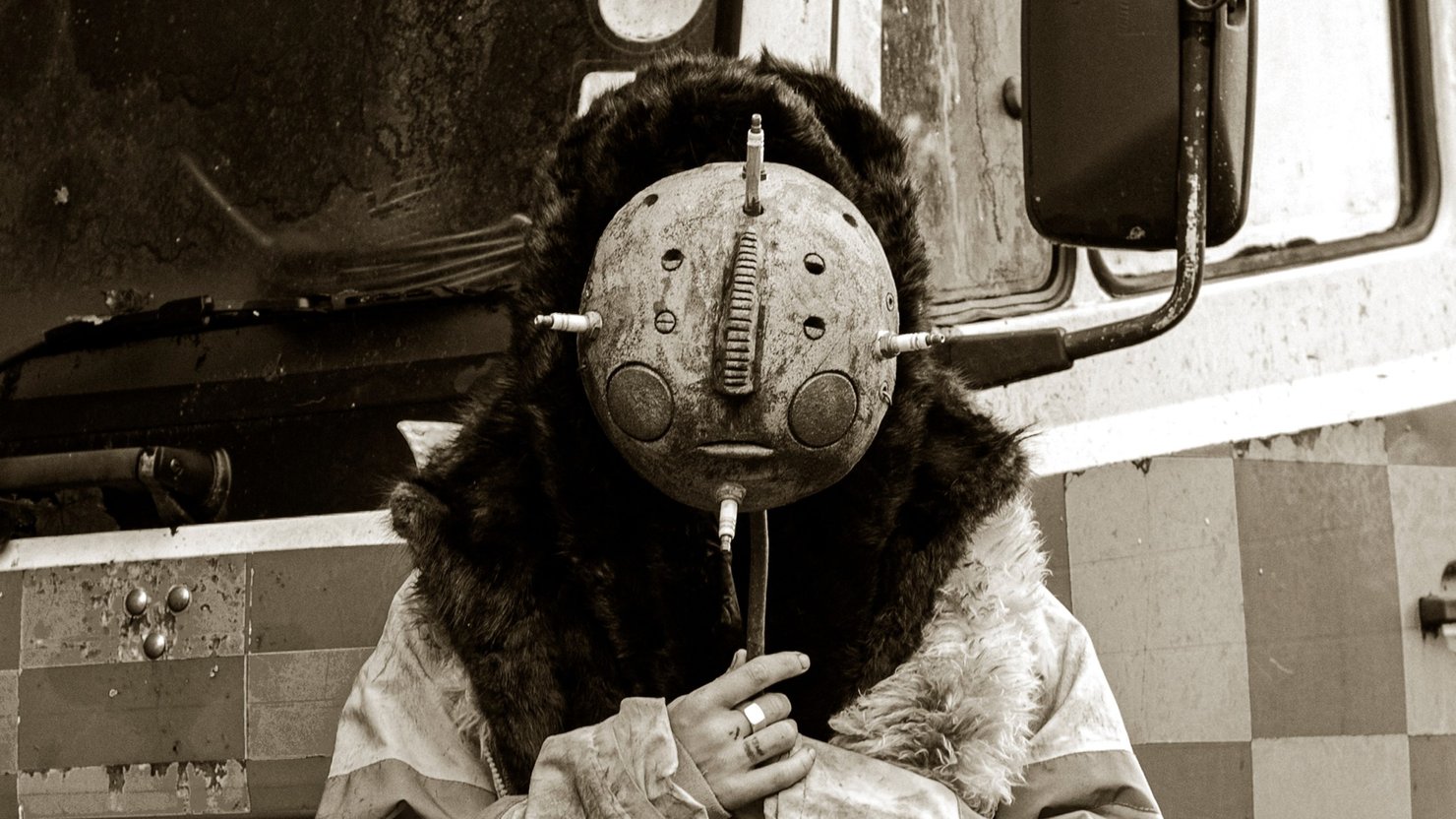 Photograph showing a still from a film, with a person covering their face with a mask
