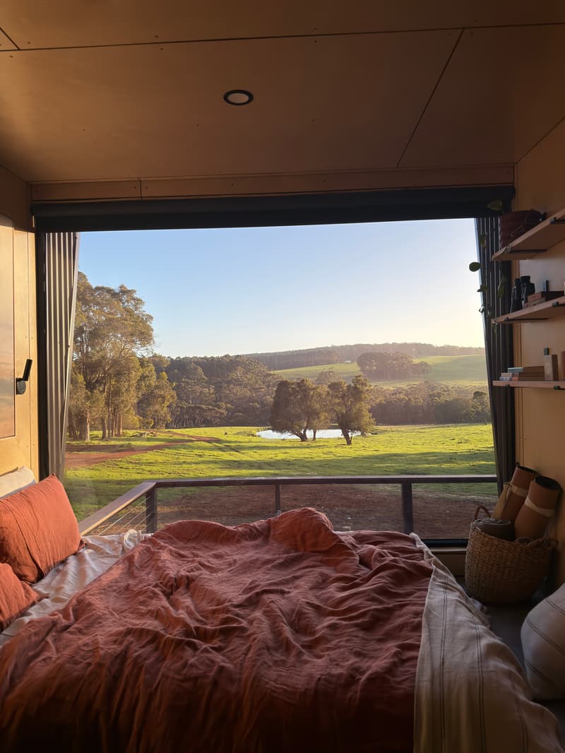 Bed facing an open wall onto paddocks at sunrise, tiny cabin, Denmark WA