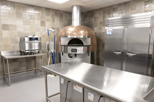 Commercial kitchen with a copper-domed pizza oven, stainless steel prep table, double-door refrigerator, and countertop cooking equipment against tiled walls.