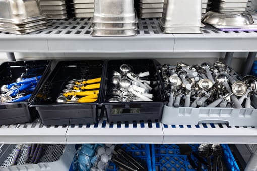 Organized shelving with bins of kitchen utensils, including scoops, ladles, tongs, and portioning tools in a commercial kitchen storage area.