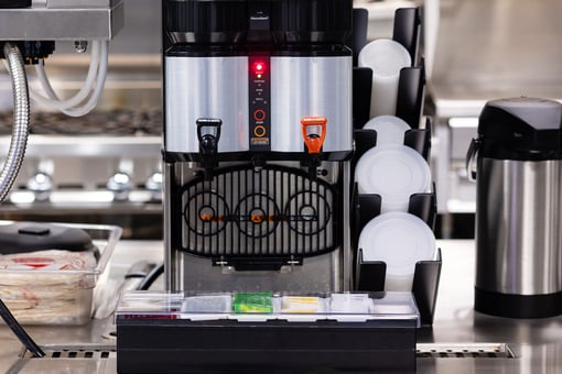Commercial beverage dispenser with dual spigots, stacked cups, and condiment tray on a stainless steel countertop in a foodservice setting.