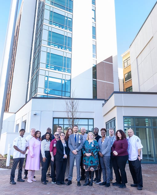 Group of hospital foodservice staff and administrators standing outside a modern medical facility building, posing together in front of a glass tower entrance.