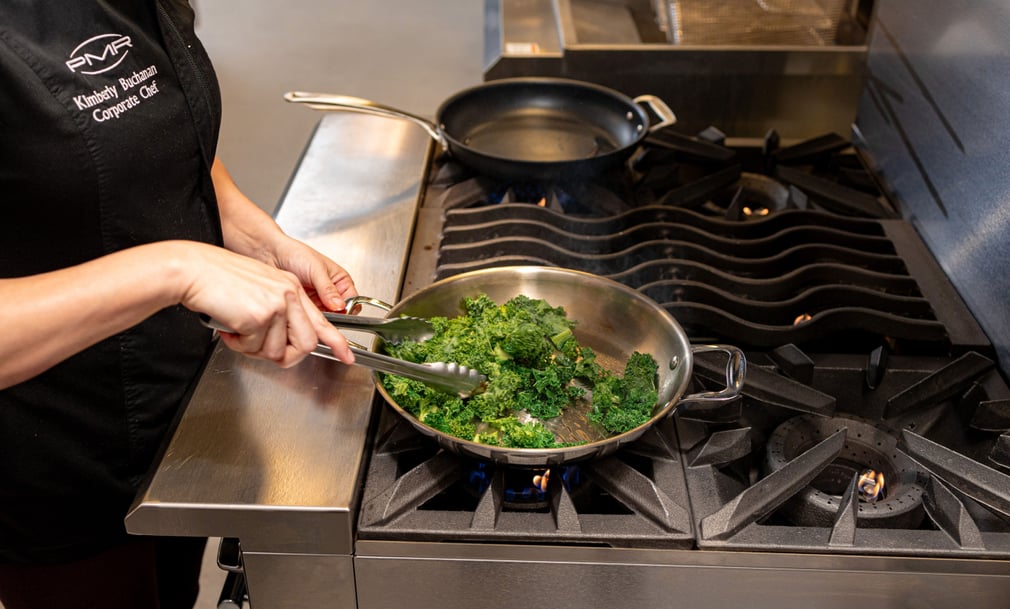 A chef cooking leafy greens in a stainless steel skillet on a commercial gas range. The chef is using tongs to stir the greens, and another pan is on a nearby burner in a professional kitchen setting.