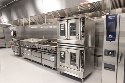 Commercial kitchen with large stainless steel tilting kettles and braising pans, integrated faucets, floor drains, and overhead ventilation hood.