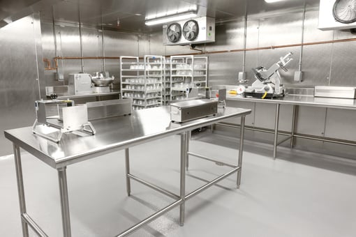Cold prep room with stainless steel tables, meat slicer, food processing equipment, and shelving units holding containers in a refrigerated commercial kitchen space.