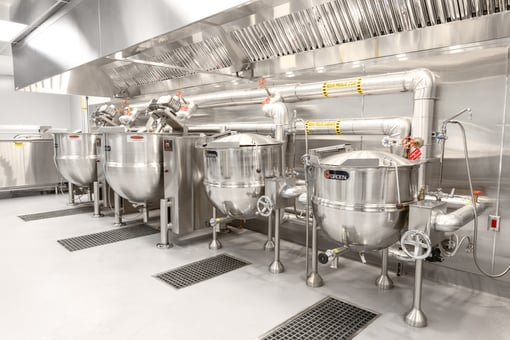 Row of large stainless steel steam kettles with plumbing connections and floor drains beneath a commercial ventilation hood in an industrial kitchen.