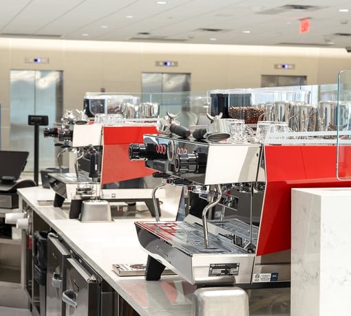Row of commercial espresso machines with grinders and cups on a countertop in a café or cafeteria beverage station.