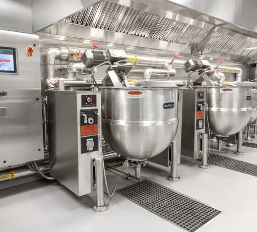 Large stainless steel steam kettle with control panel and piping connections beneath a commercial ventilation hood in an industrial kitchen.