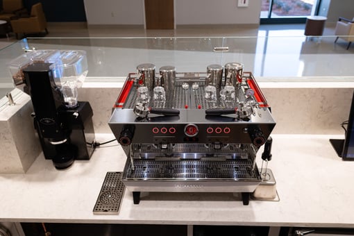 Commercial espresso machine with multiple group heads and glassware on top, positioned on a countertop next to a coffee grinder in a café-style setup.