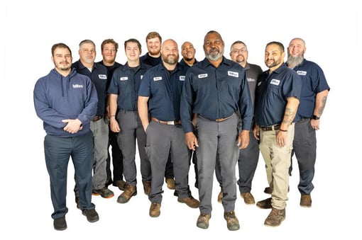 Group of eleven uniformed technicians standing together in a studio, wearing matching dark blue work shirts and pants with name patches, facing the camera in a staggered formation against a plain white background.