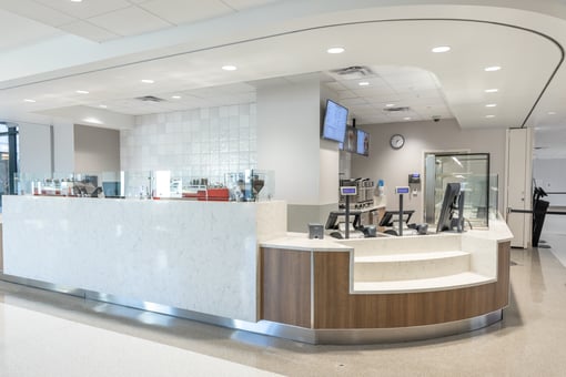 Modern cafeteria service counter with marble-style front, POS terminals, overhead menu screens, and glass sneeze guards in a clean, contemporary dining area.