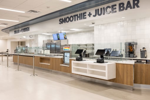 Cafeteria service line with "Smoothie + Juice Bar" and "Salad + Garden" stations, featuring glass sneeze guards, POS terminals, menu screens, and beverage equipment in a modern dining area.