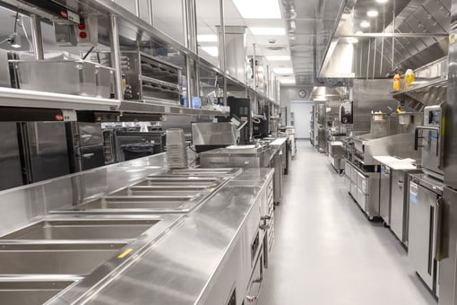 Long stainless steel commercial kitchen line with prep wells, cooking equipment, overhead shelving, and ventilation hoods in a modern foodservice facility.