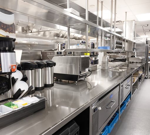 Stainless steel commercial kitchen workspace with countertop appliances, beverage dispensers, storage drawers, and shelving in a clean, industrial food prep area.