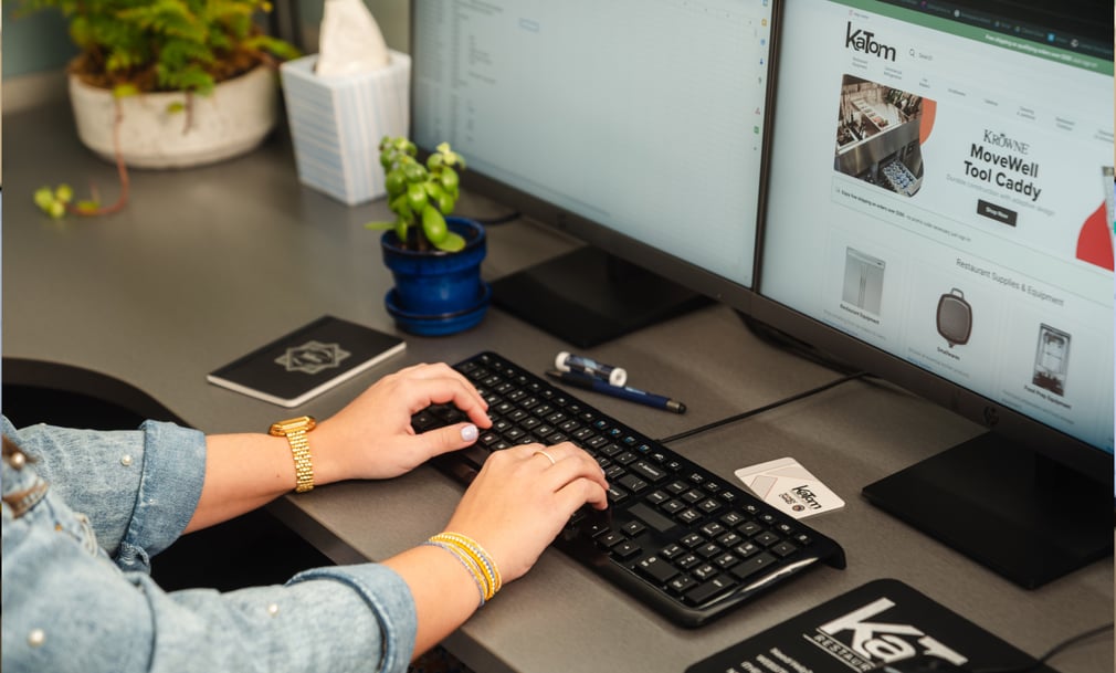Close-up of a person typing on a keyboard at a modern desk with dual monitors. One screen shows the KaTom website, while the other displays a document or spreadsheet. A small plant, notebook, and office supplies are also on the desk.