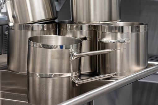 Stacked stainless steel stock pots and saucepans with long handles arranged on a commercial kitchen prep shelf.