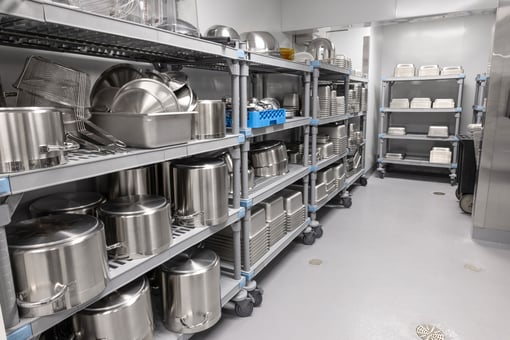 Storage room with organized shelving holding stainless steel pots, pans, trays, and kitchen equipment on wheeled racks in a commercial kitchen.