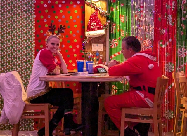 Reindeer-antlered woman shares a quiet table with a full-suit Santa amid green-and-red foil fringe