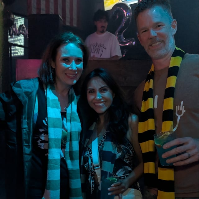 Three wand-toting friends in house scarves and robes beam over potion glasses at a dim Wizards Night Out tavern.