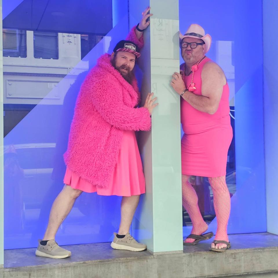 Two men in hot pink dresses, fuzzy coat and cowboy hat strike playful poses against a blue wall