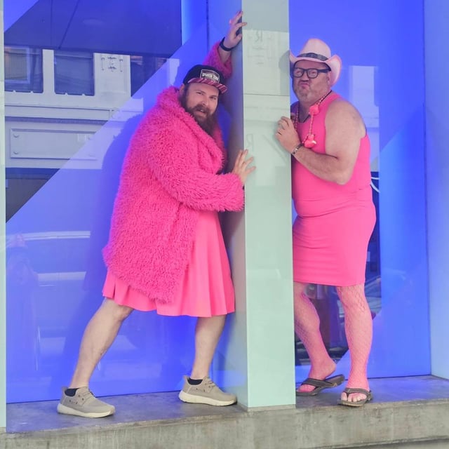 Two men in hot pink dresses, fuzzy coat and cowboy hat strike playful poses against a blue wall