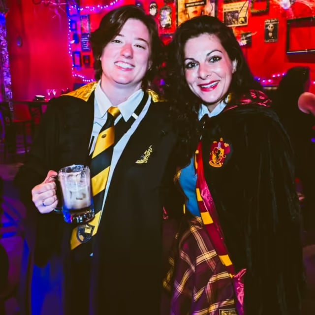 Hufflepuff and Gryffindor witches in full robes share a frothy butterbeer mug on the wizard crawl