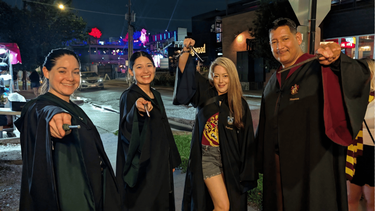 Four robed wizards-in-training raise wands toward the camera on a neon-lit street between Wizards Night Out stops.