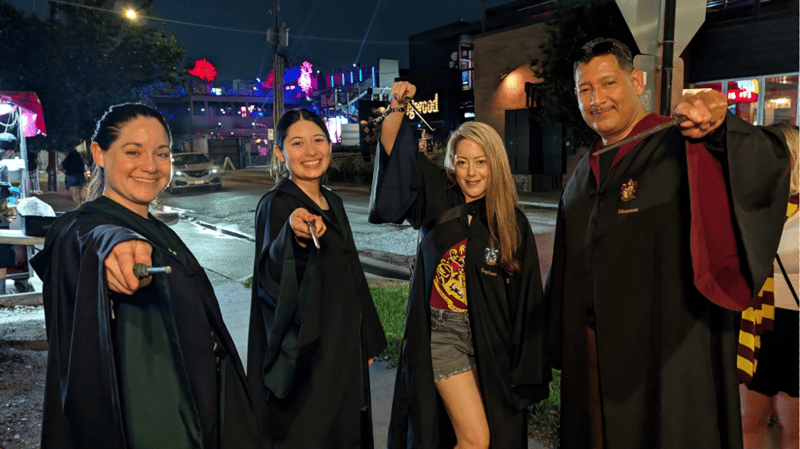 Four robed wizards-in-training raise wands on a neon-lit street between Wizards Night Out stops