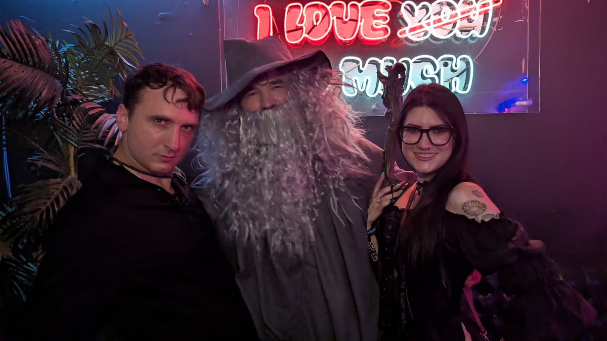 Trio flanks the long-bearded wizard character with staff beneath a glowing neon sign on the wizard crawl