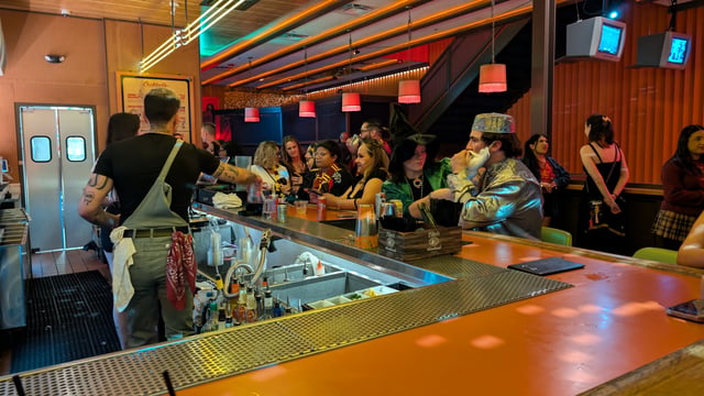 Costumed wizard crawl crowd lines the bar as a muggle bartender mixes themed brews for robed witches and wizards along the rail.
