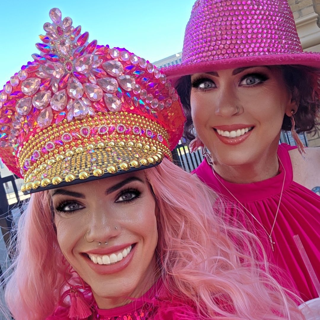 Close selfie of two women in jeweled pink captain hat and rhinestone fedora with pink wigs