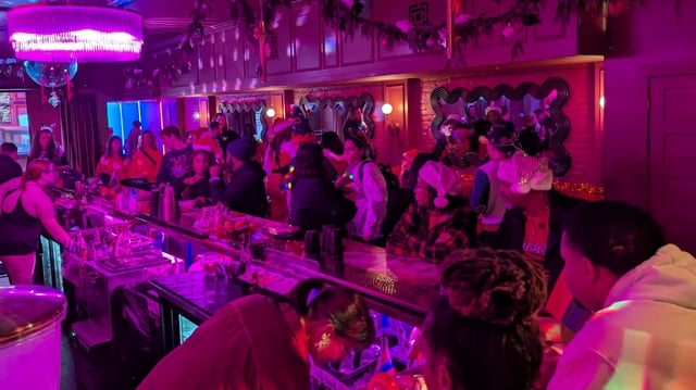 Pink-and-purple lit bar packed with elf-hatted revelers under a chandelier and garland-draped mirror