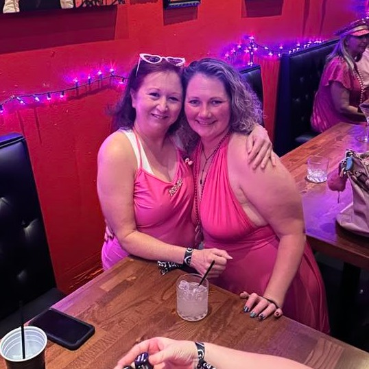 Two women in pink dresses snuggle in a red-lit bar booth with a cocktail and string lights