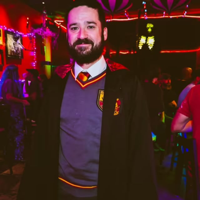 Bearded wizard-in-training in full Gryffindor robes and house tie grins inside a red-lit Wizards Night Out stop
