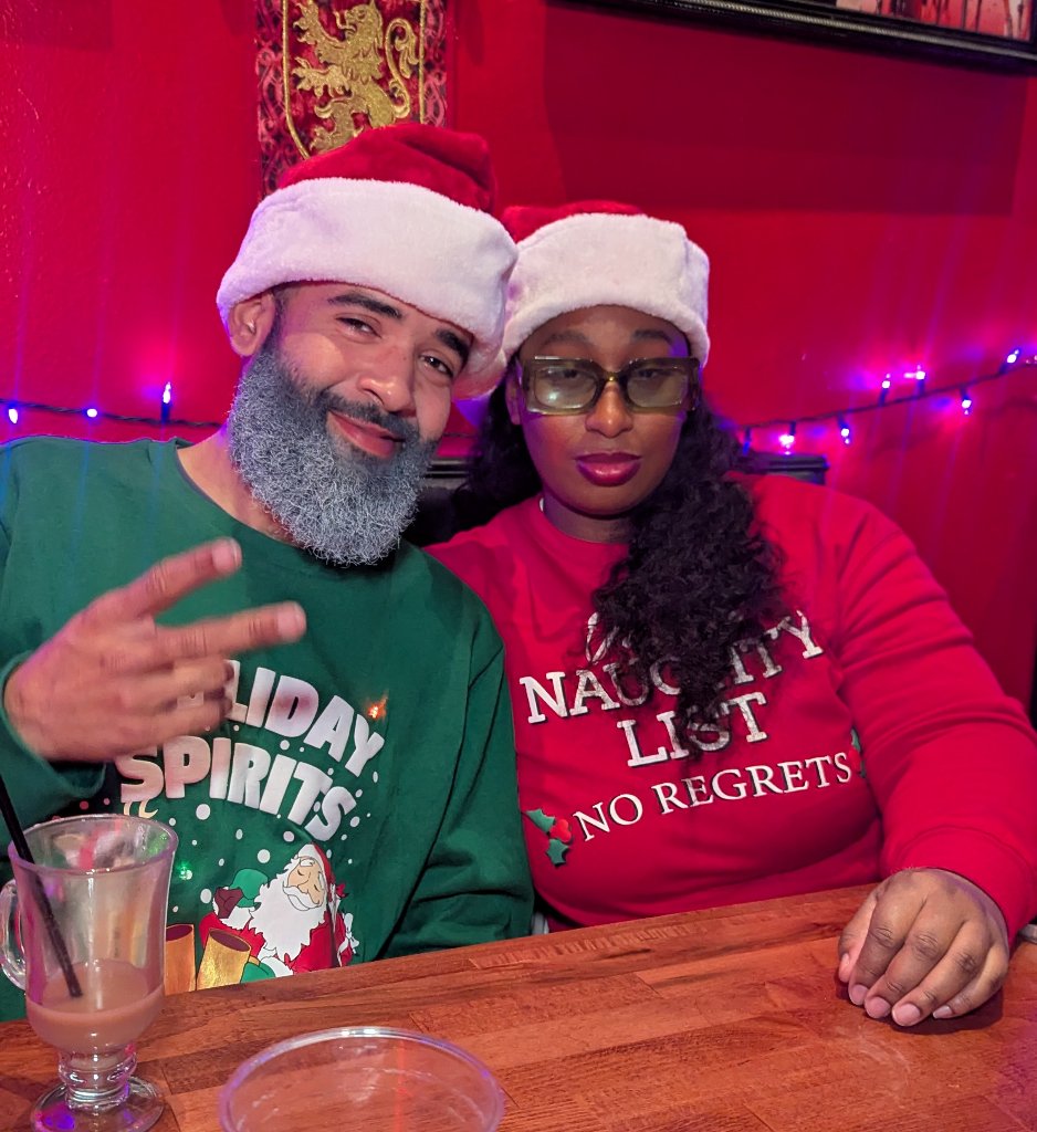Couple in Santa hats and matching Holiday Spirits and Naughty List sweaters cozy up in a red booth