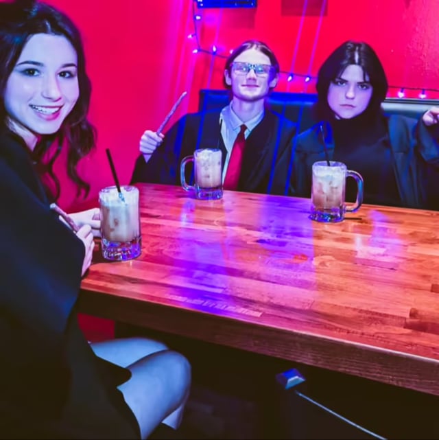 Three Hogwarts-style students brandish a wand over butterbeer mugs at a Wizards Night Out booth