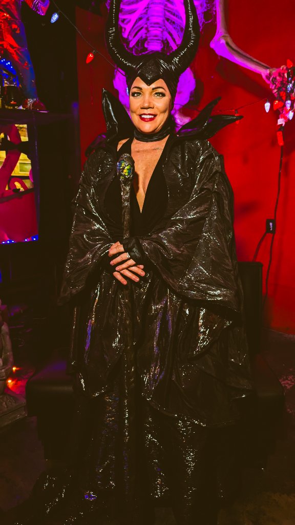 Maleficent in horned headpiece and glossy black cloak smiles before a skeleton-and-fog spooky bar backdrop