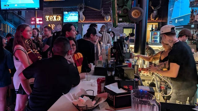 Wizard crawl crowd in house scarves waits as a tattooed muggle bartender mixes potions beneath a neon Texas sign.