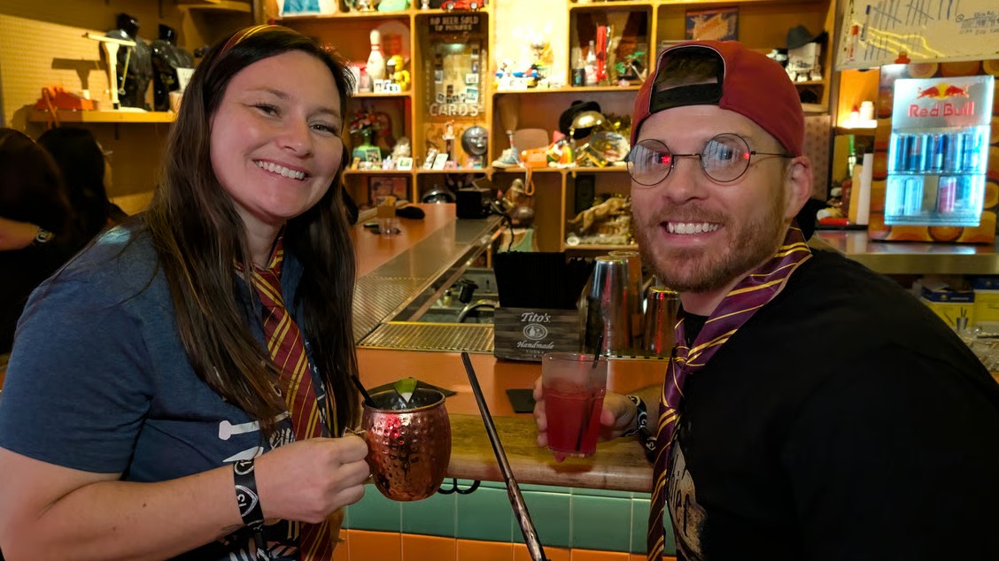 House-tied wizard crawl pair clink a copper mug and red potion glass, wand laid across the bar at a quirky stop.