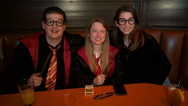 Three wand-wielding wizards in round Potter glasses, robes, and house ties hold court at a Wizards Night Out booth.