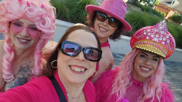 Four women in pink wigs, sequined hats and hot pink tops squeeze into a bright outdoor selfie