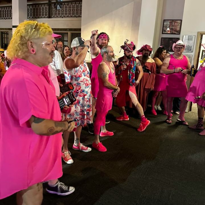 Crowd in pink dresses, wigs and feathered hats gather inside a venue